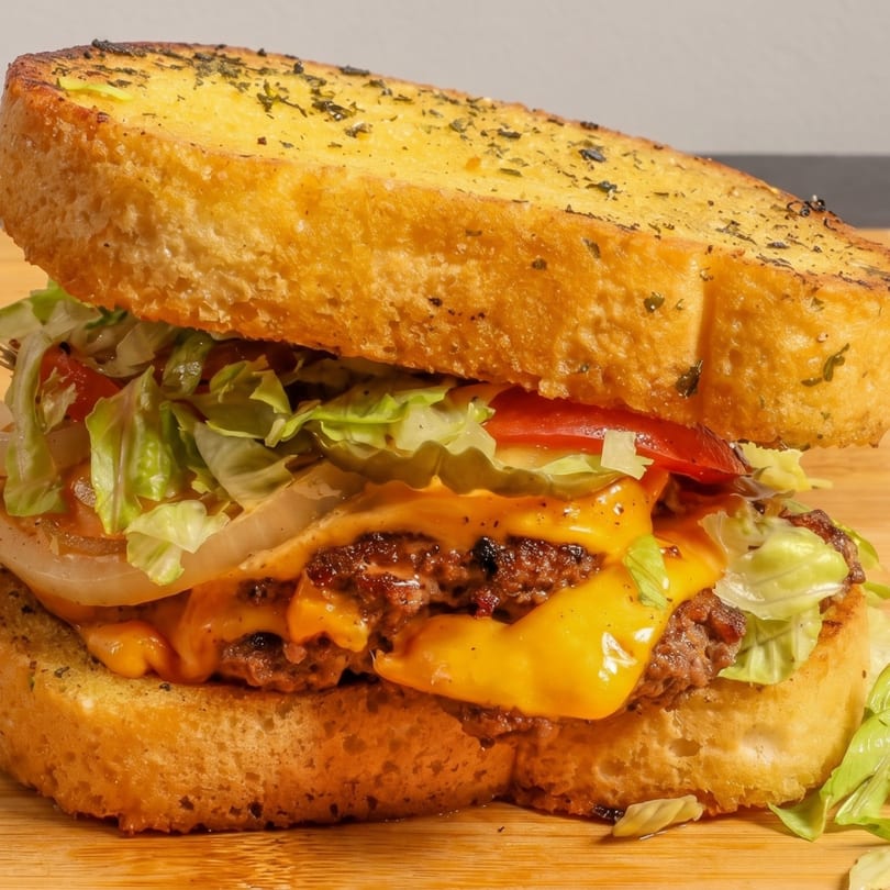 Close-up of a cheeseburger on garlic bread, with lettuce, tomato, and onion on a wooden board.