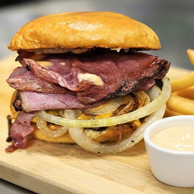 A pastrami sandwich with grilled onions and sauce on a wooden board with fries and dipping sauce.